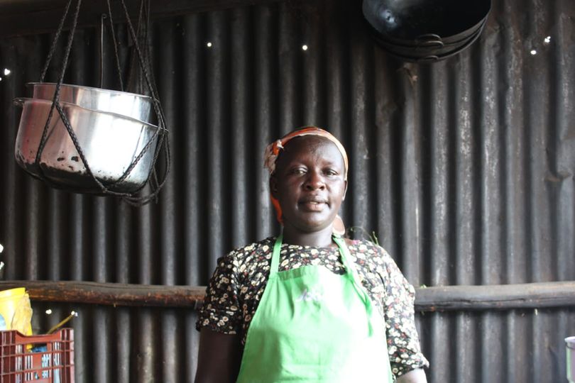 A head and shoulders photograph of Macrine taken in her eatery, with corrugated iron and pots in the background. Macrine wears a floral blouse and green apron.